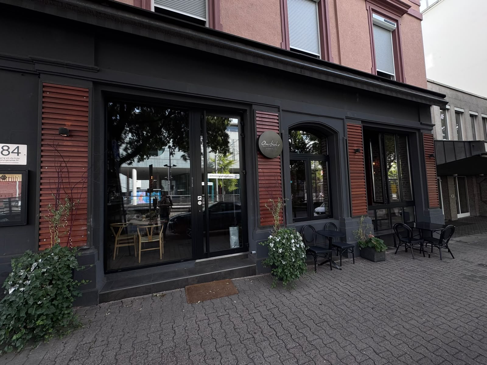Die Oinotheke von der Straße: dunkle Fassade mit rotbraunen Läden und Tischen davor
